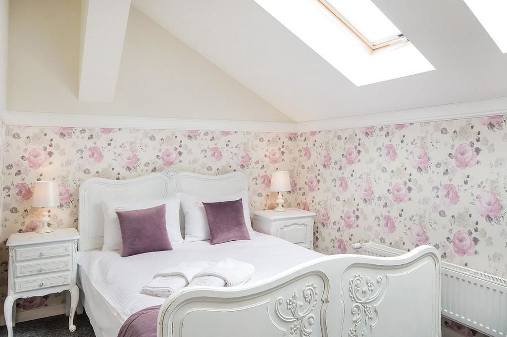 A bedroom with a white bed, floral wallpaper, two nightstands with lamps, and a skylight.