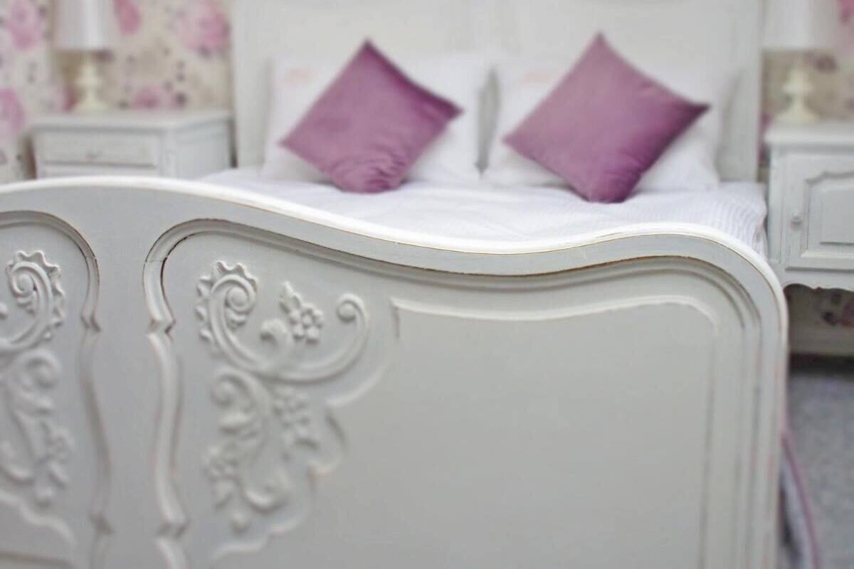 A white bed with ornate carvings and two purple pillows.