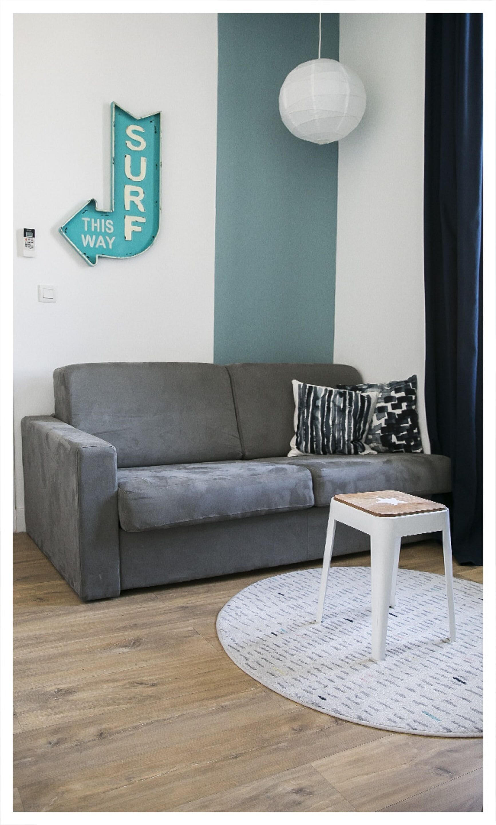 A grey sofa with patterned cushions, a white side table, and a round rug on a wooden floor. A sign with the word "SURF" is mounted on the wall.