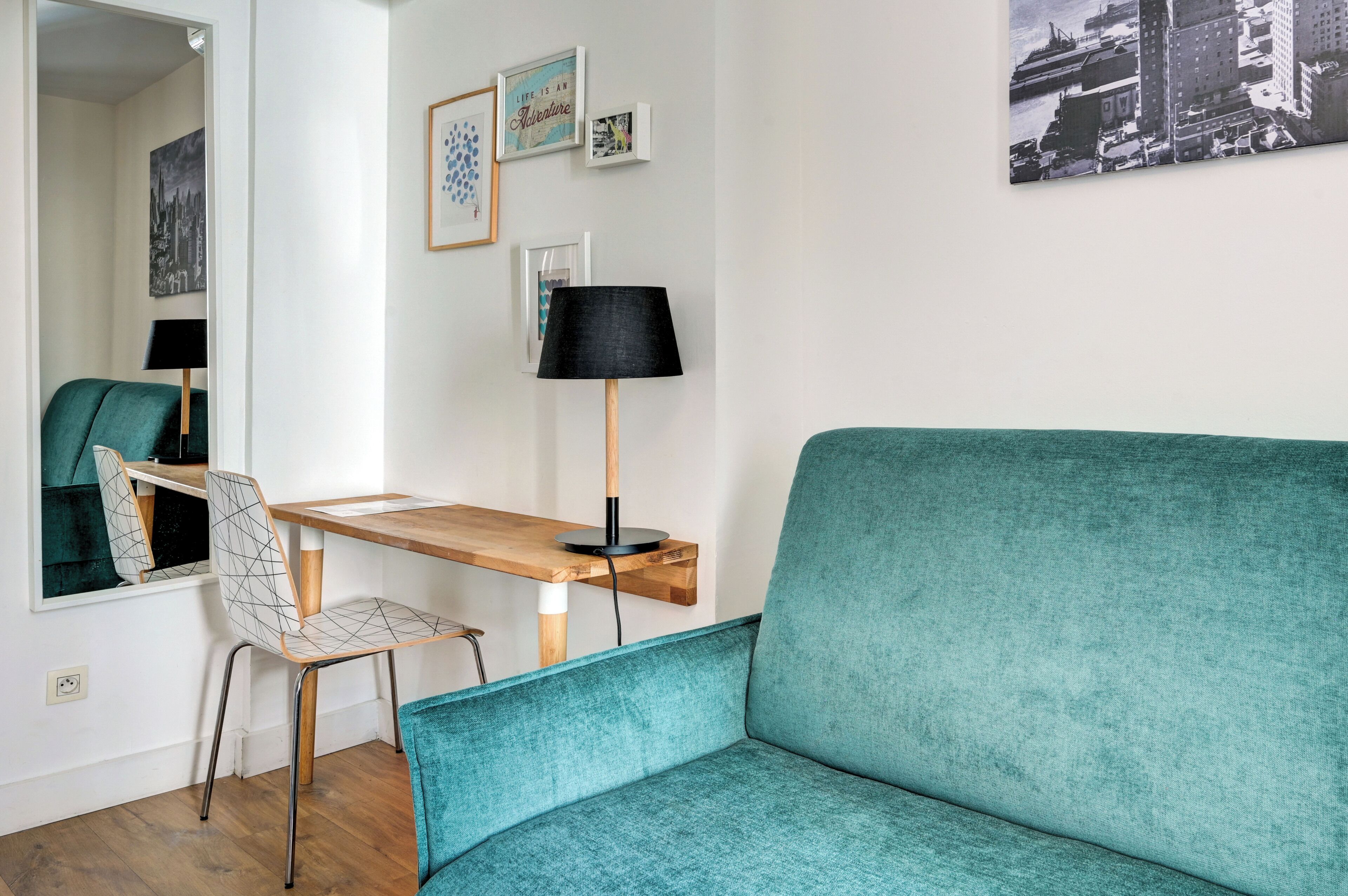 A room with a teal sofa, a wooden desk with a lamp, a chair, and a wall with framed pictures.