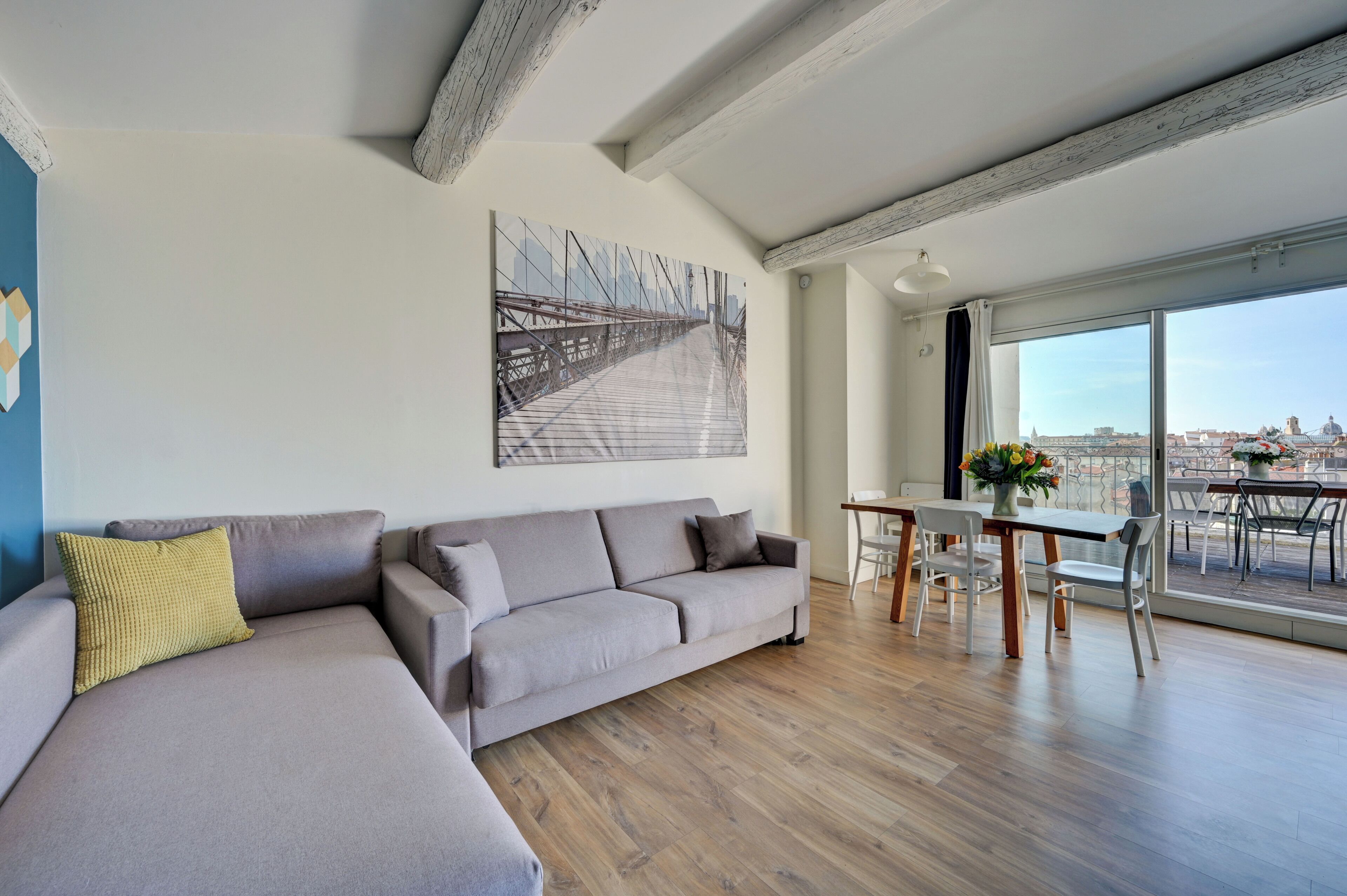 A modern living room with a grey sofa, wooden flooring, a dining area with a table and chairs, a large window with a city view, and a painting on the wall.