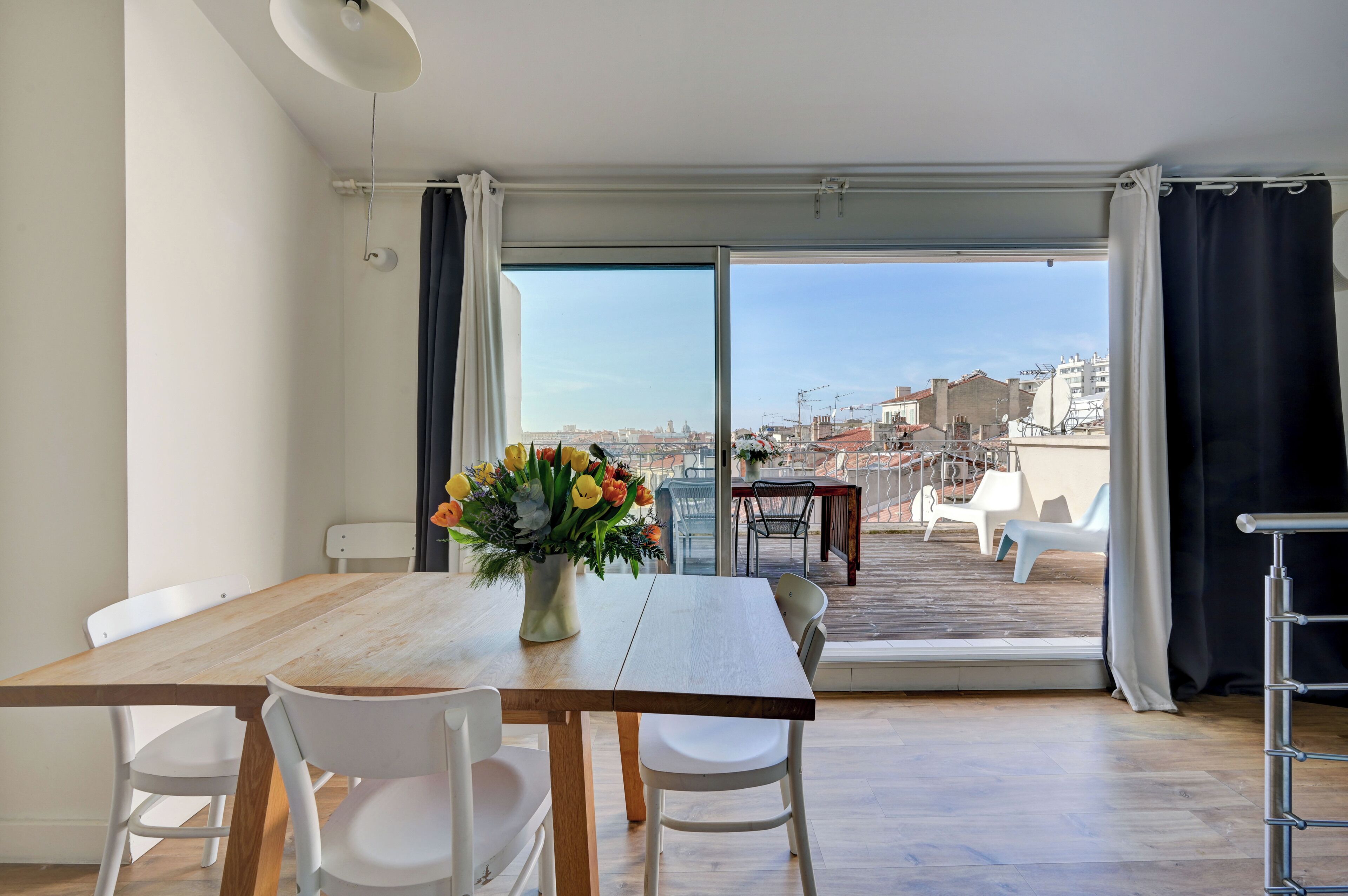A modern dining area with a wooden table and white chairs, a vase of flowers, and a view of a cityscape through large glass doors.