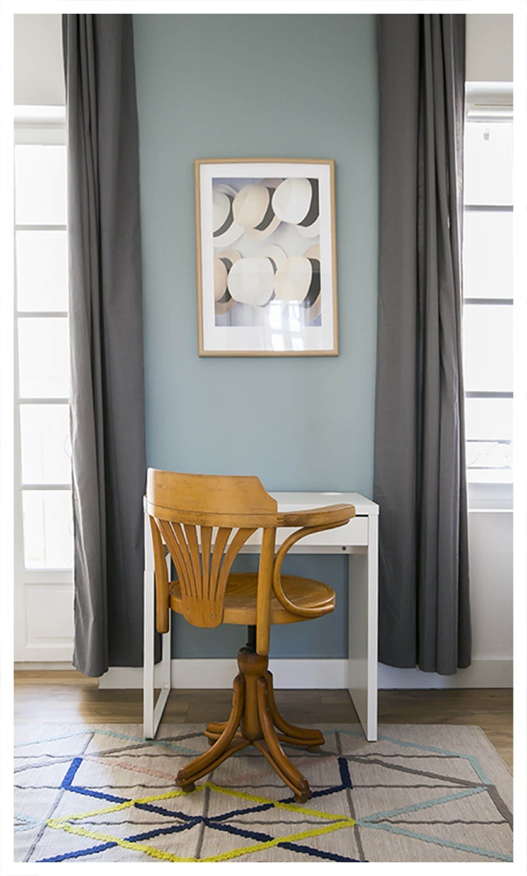 A room with a wooden chair, a white desk, and a framed artwork on the wall.