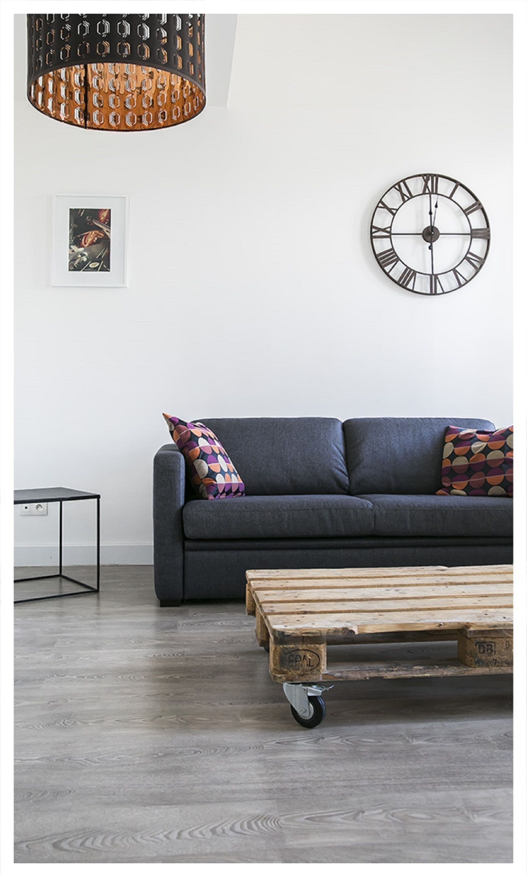 A modern living room with a dark grey sofa, colorful patterned pillows, a wooden coffee table with wheels, a black metal clock, and a pendant light with a geometric design.