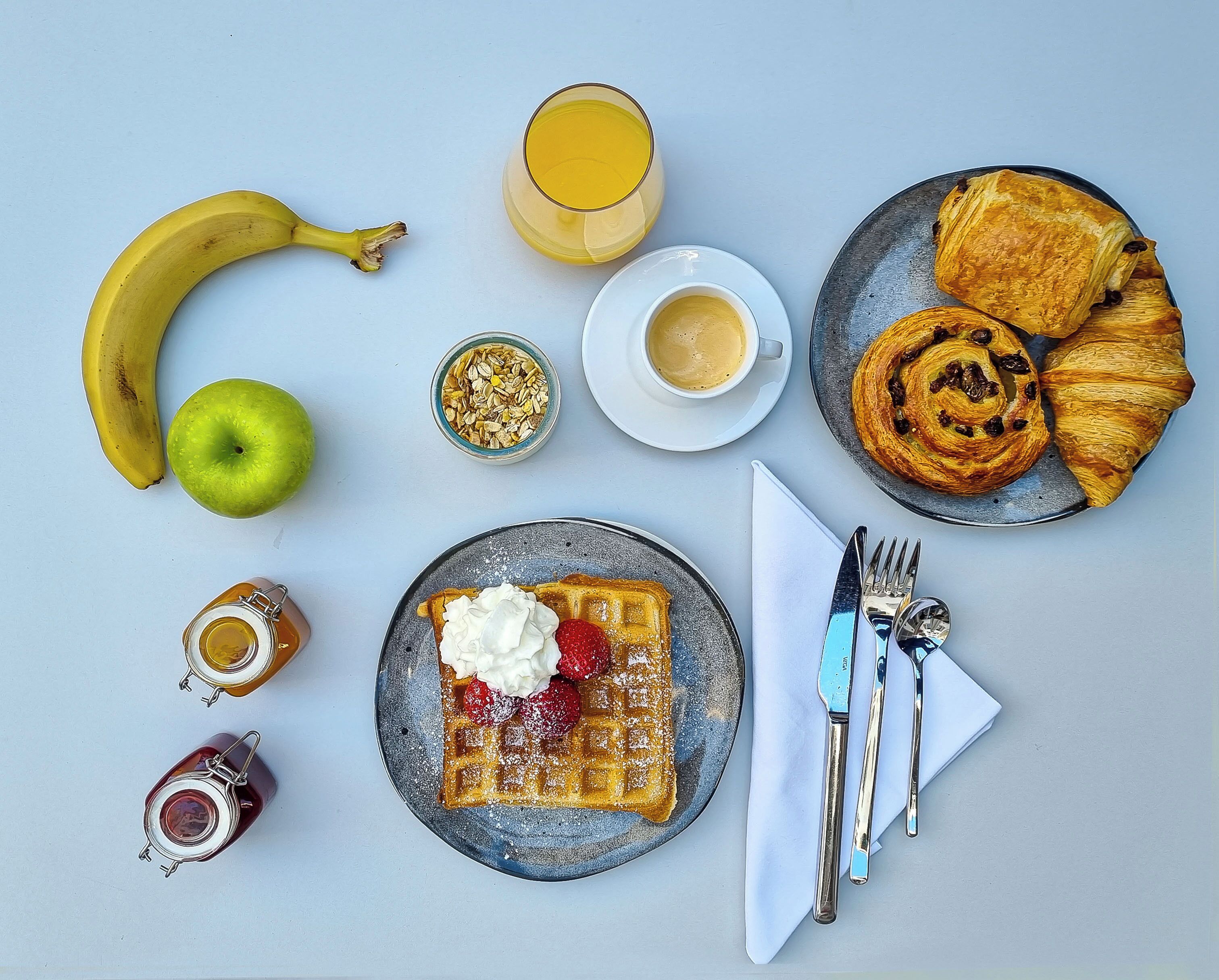 A breakfast setting with a banana, apple, waffles with whipped cream and strawberries, croissants, a glass of orange juice, a cup of coffee, and a jar of jam.