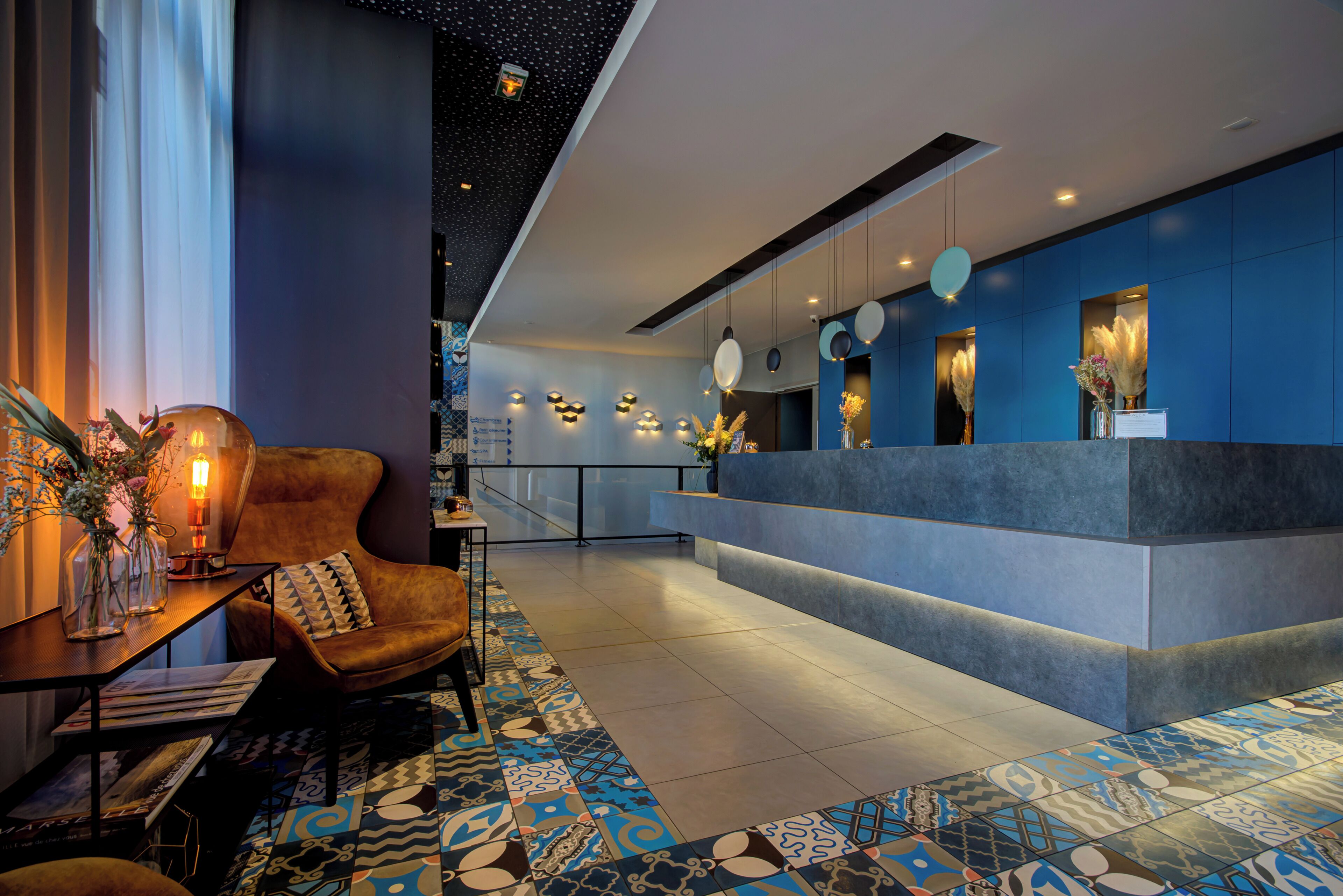 A modern hotel lobby with a reception desk, seating area, and patterned floor tiles.
