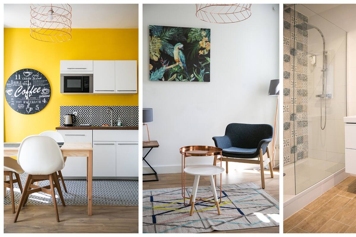A modern kitchen with a yellow accent wall, a coffee-themed wall art, a dining area with a white chair, and a patterned rug.
