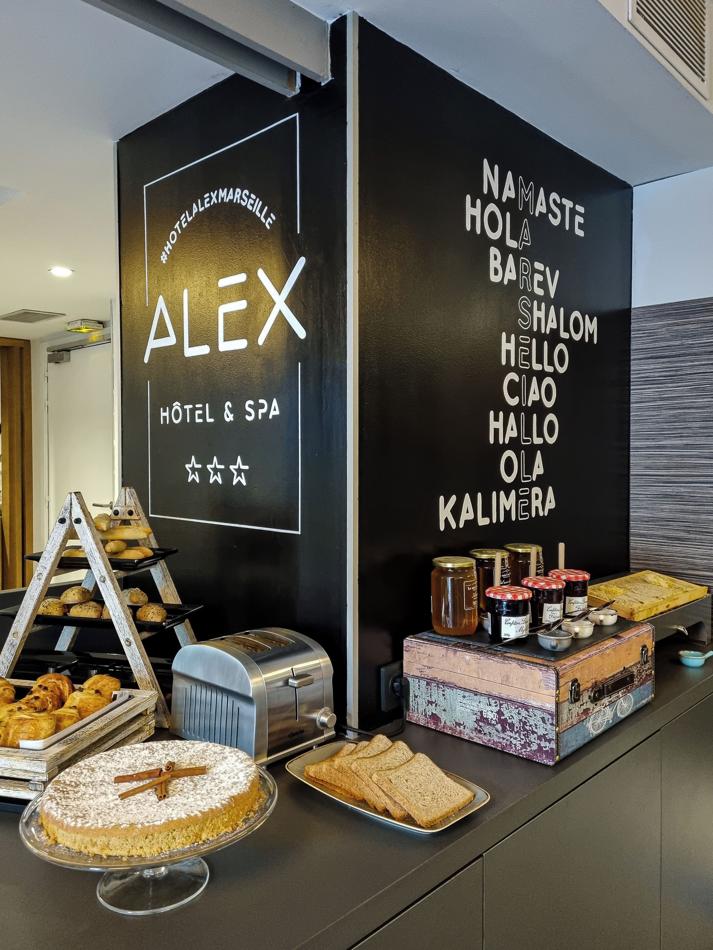 Zona de recepción de un hotel con un cartel que dice 'HOTEL & SPA ALEX' y una pared mostrando saludos en varios idiomas.