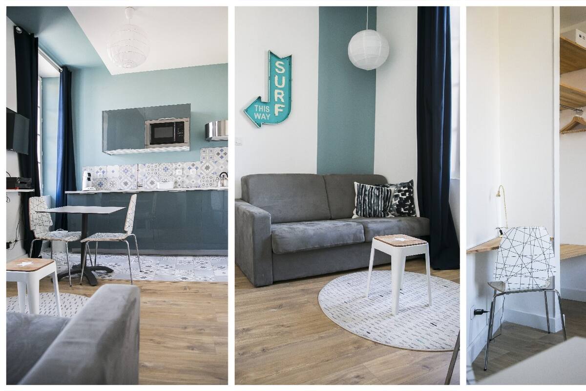 A modern living room with a grey sofa, a small round table, and a white chair. There is a kitchen area with a microwave and a sink. A sign on the wall reads "SURF THIS WAY".