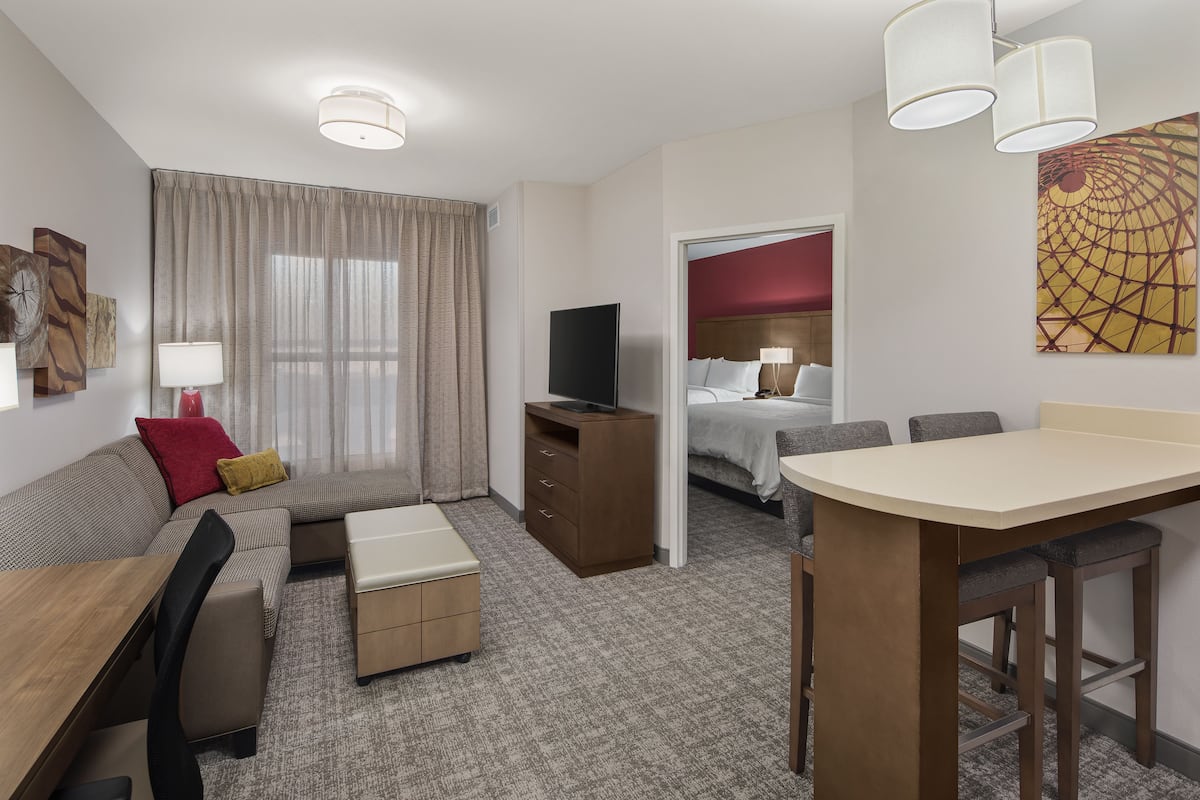 A hotel room with a sofa, a small table, a TV, a desk, and a chair. There is a window with curtains, a painting on the wall, and a door leading to another room.
