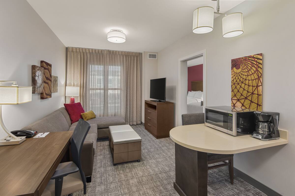 A hotel room with a sofa, a dining table, a coffee maker, a TV, and artwork on the wall.