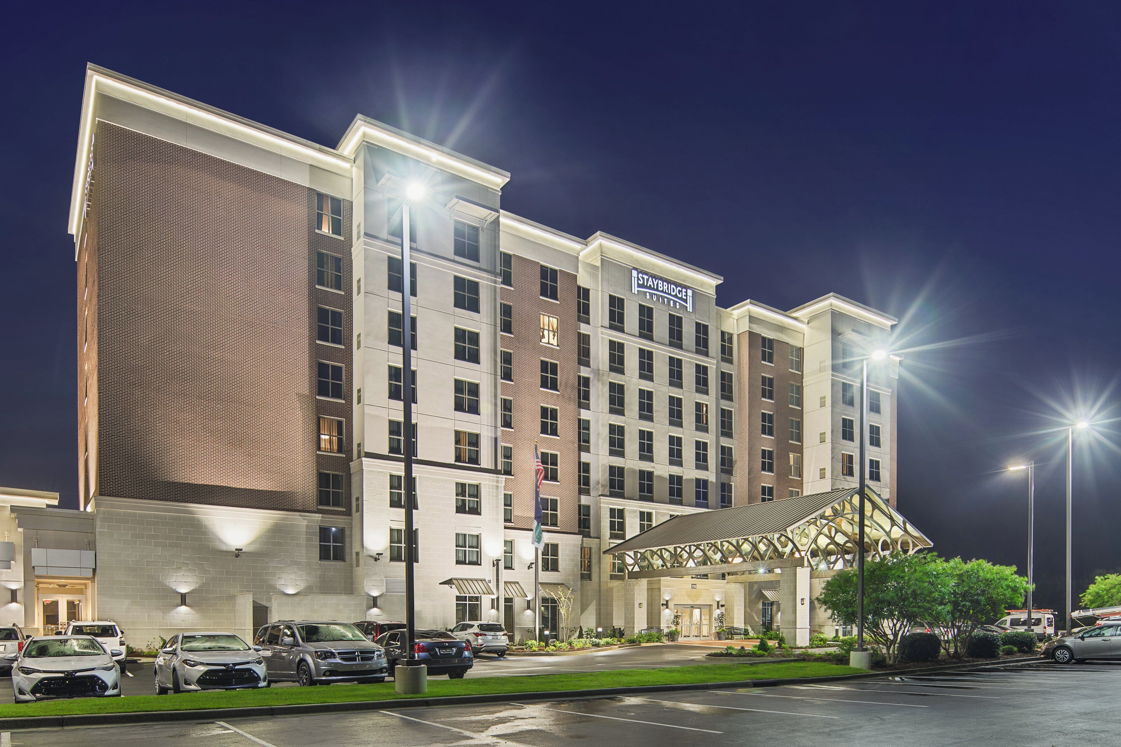 A multi-story hotel with a prominent Staybridge Suites sign, a covered entrance, and a parking lot with several cars.