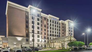 A multi-story hotel with a prominent Staybridge Suites sign, a covered entrance, and a parking lot with several cars.