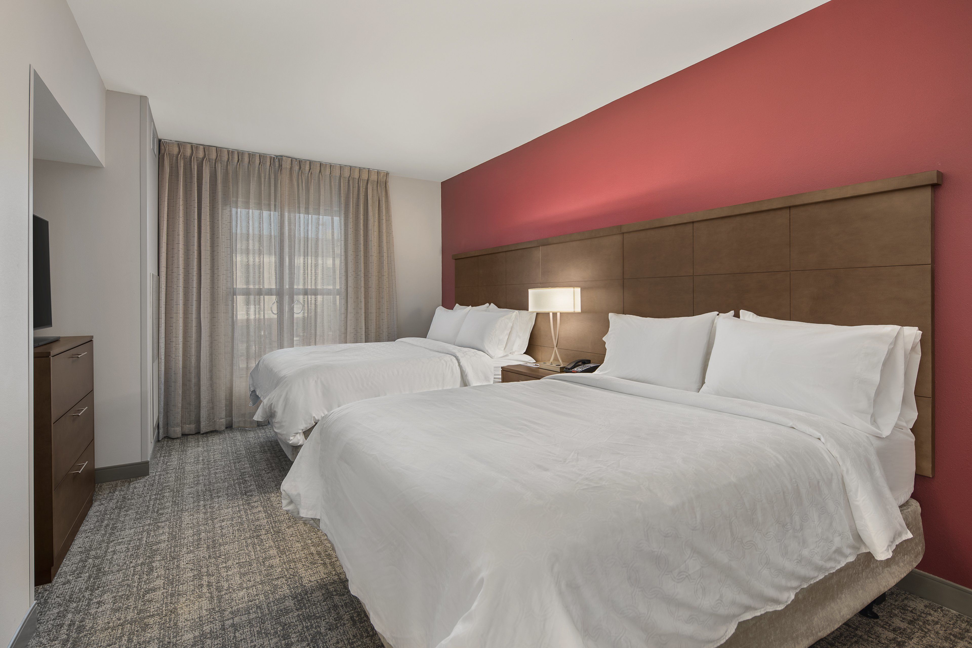 A hotel room with two beds, a wooden headboard, a bedside table with a lamp, and a window with curtains.
