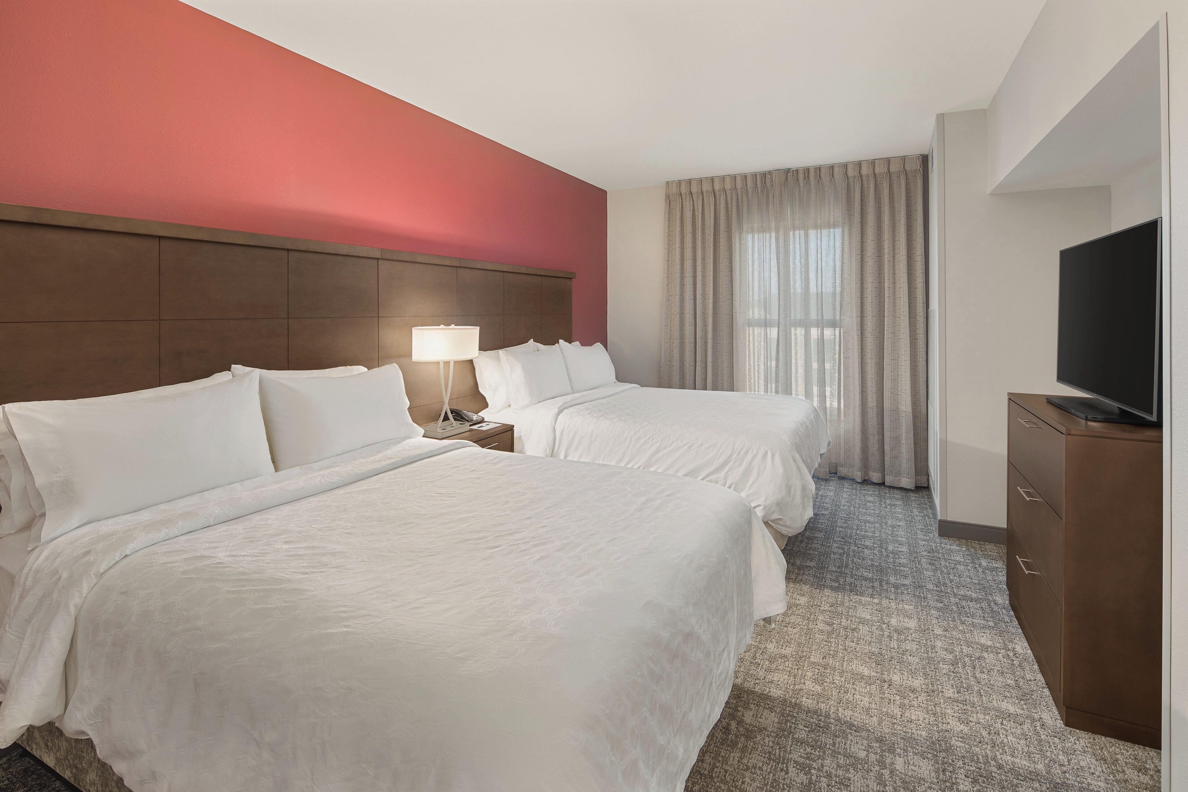 A hotel room with two beds, a nightstand with a lamp, a television on a cabinet, and a large window with curtains.