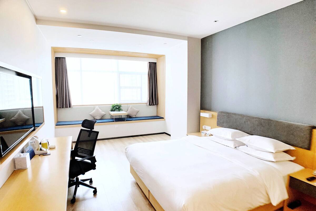 A hotel room with a bed, a desk with a chair, a TV, and a window with curtains.