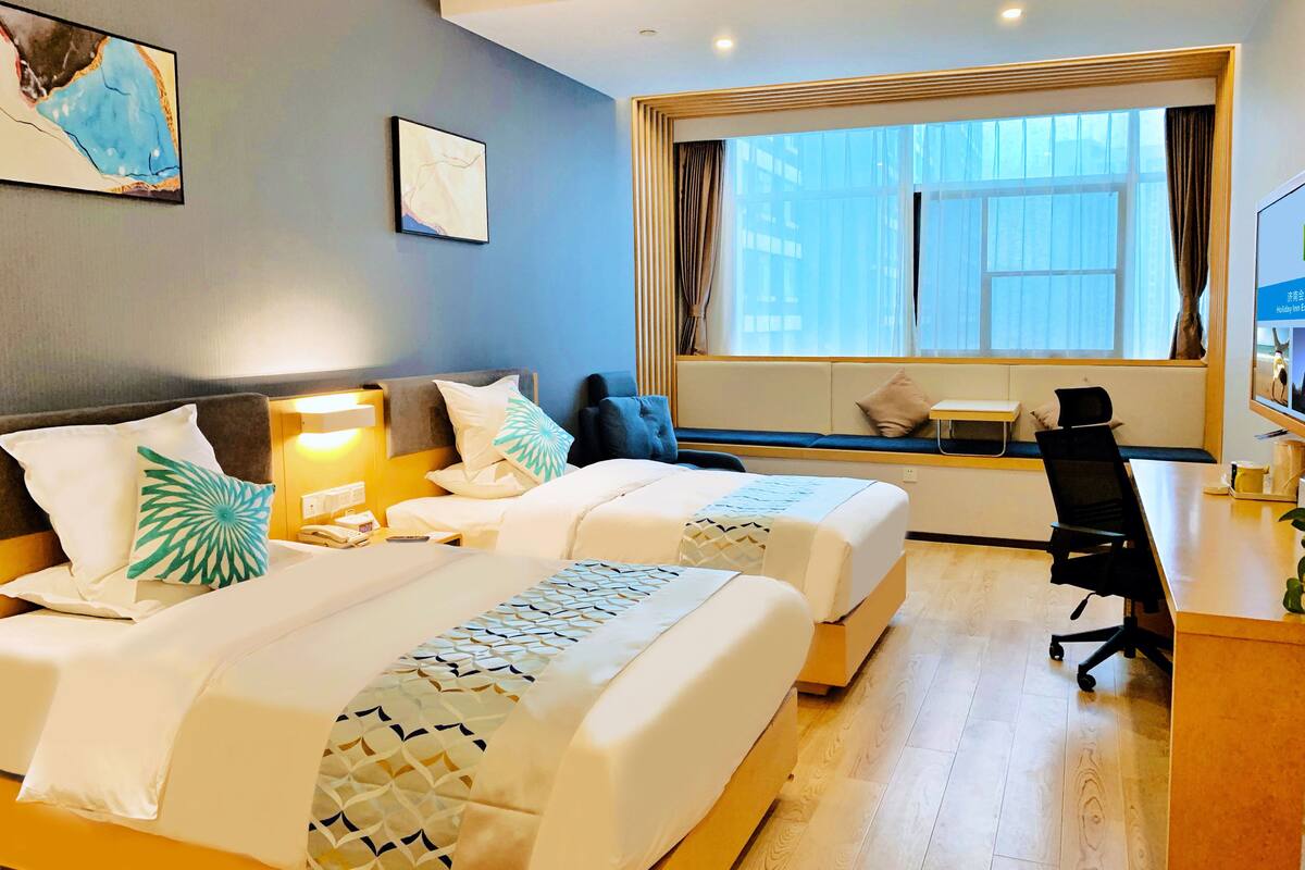 A hotel room with two beds, a desk, a chair, and a large window with curtains.