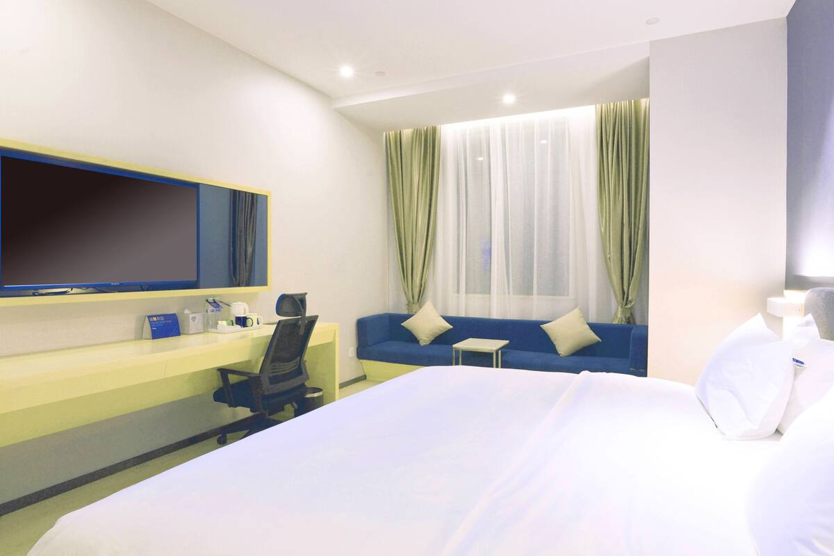 A hotel room with a bed, a desk with a computer, a chair, a TV mounted on the wall, and a window with curtains.