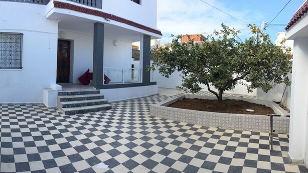 Un bâtiment blanc avec un sol à carreaux, un petit porche avec des marches et un arbre dans la cour.