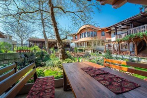 An outdoor seating area with wooden benches and tables, surrounded by lush greenery and a stone wall. - Hotel Izvora (Kranevo)