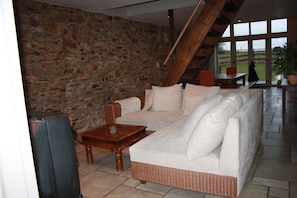 A cozy living room with a stone wall, a wicker sofa, a wooden coffee table, and a view of the outdoors through large windows. - Dry Le Nou Menatye (Manhay)