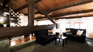 A spacious room with a high ceiling, wooden beams, a black sofa set, a coffee table, and a checkered floor pattern.