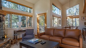 A spacious living room with a large brown leather sofa, a wooden coffee table, and a stone fireplace. Large windows provide ample natural light and views of the surrounding forest. - Bridgepoint Condo 8 - Freshly Updated, Baldy Views, Walk to Downtown & Lifts (Ketchum)