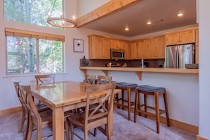 A kitchen with wooden cabinets, a stainless steel refrigerator, a microwave, and a dining area with a wooden table and chairs. - Bridgepoint Condo 8 - Freshly Updated, Baldy Views, Walk to Downtown & Lifts (Ketchum)
