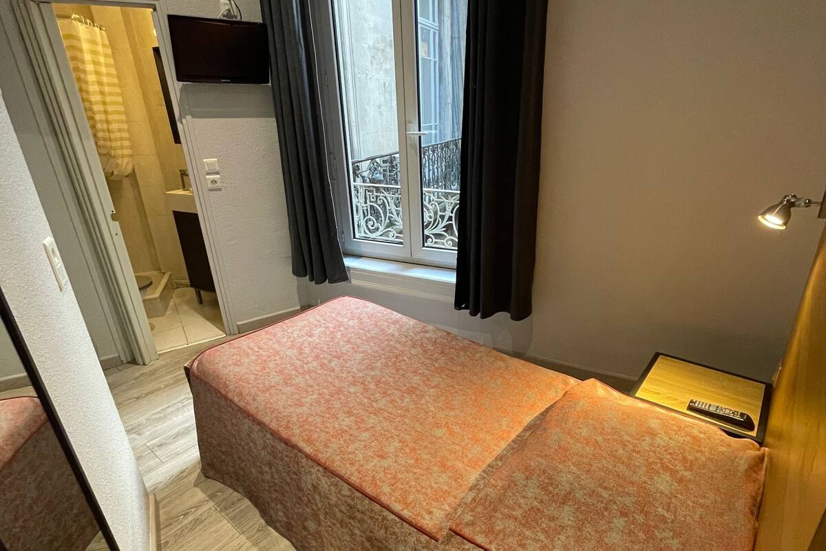 A small bedroom with a bed, a window with curtains, a bedside table, and a view of the outside balcony.