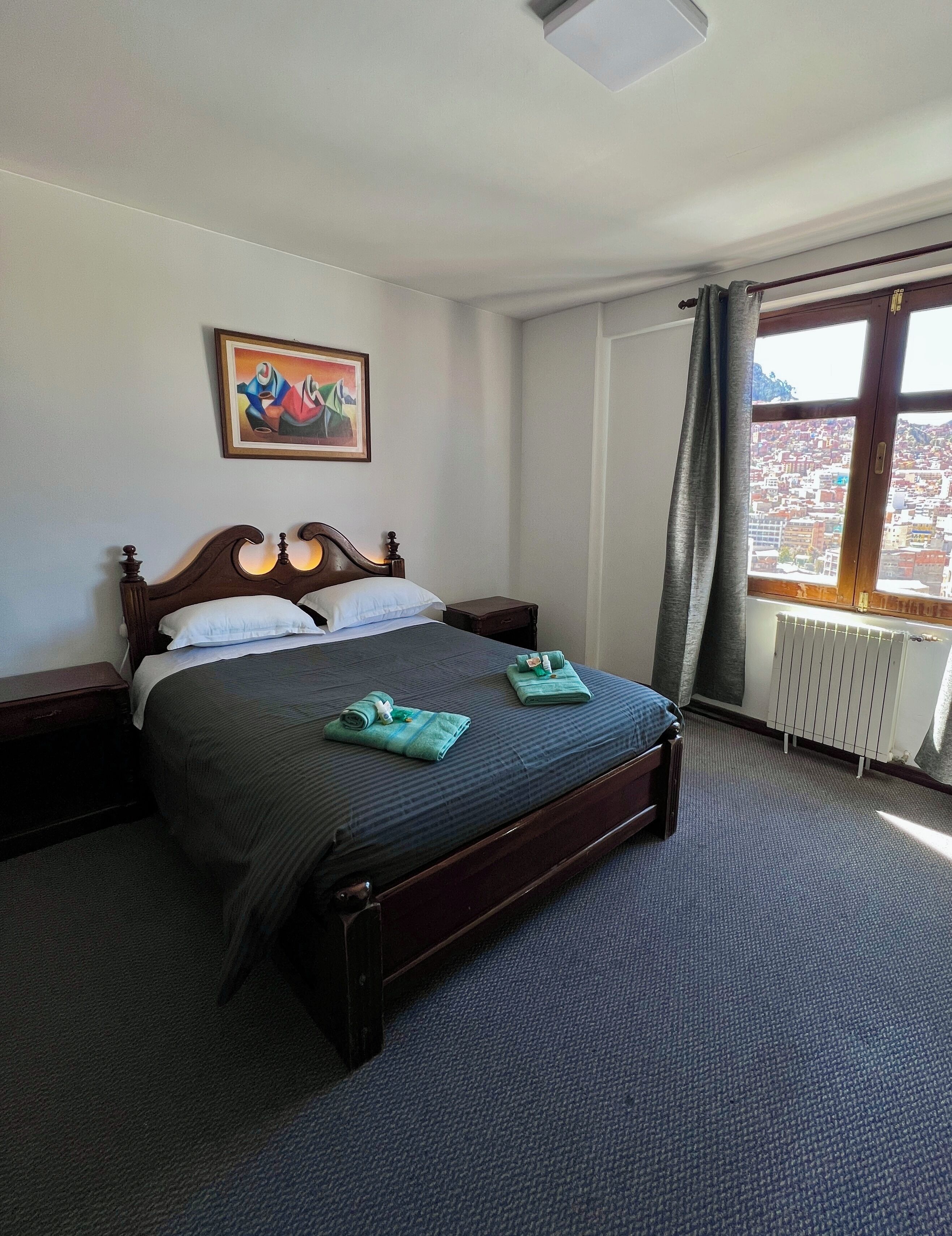 A hotel room with a bed, a painting on the wall, a window with a view, and a radiator.
