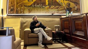 Una persona sentada en un sofá en una habitación con un mural grande en la pared que muestra diversas escenas y figuras.