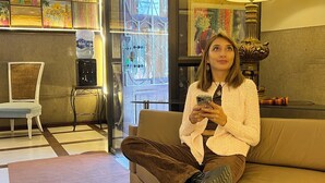 Una persona sentada en un sofá en una habitación con un ventanal grande, una lámpara y una pared decorada con varias obras de arte.
