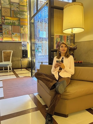 Una persona sentada en un sofá en una habitación con un ventanal grande, una lámpara y una pared decorada con varias obras de arte.