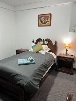 Una cama bien hecha con una colcha gris, una toalla doblada y una almohada. Se ve una mesita de noche de madera con una lámpara junto a la cama. Encima de la cama, hay un cuadro enmarcado en la pared.