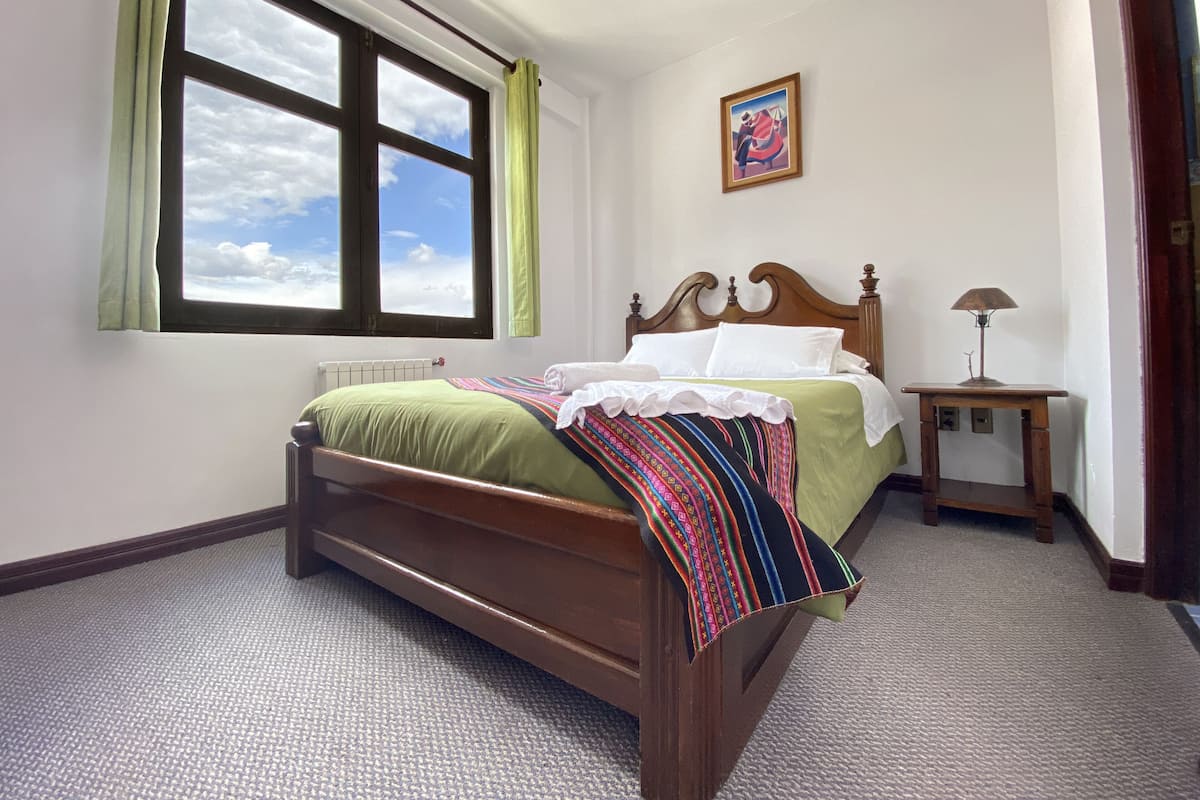 A neatly arranged bedroom with a bed, a bedside table with a lamp, a window with curtains, and a painting on the wall.