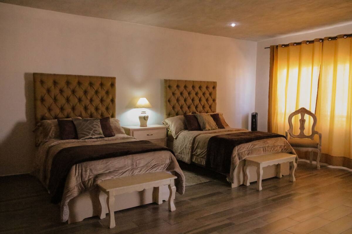 A bedroom with two beds, a nightstand with a lamp, a chair, and a bench.