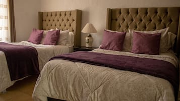 Una habitación de hotel con dos camas, cada una con cabecero capitonado, una mesita de noche con lámpara y una ventana con cortinas.