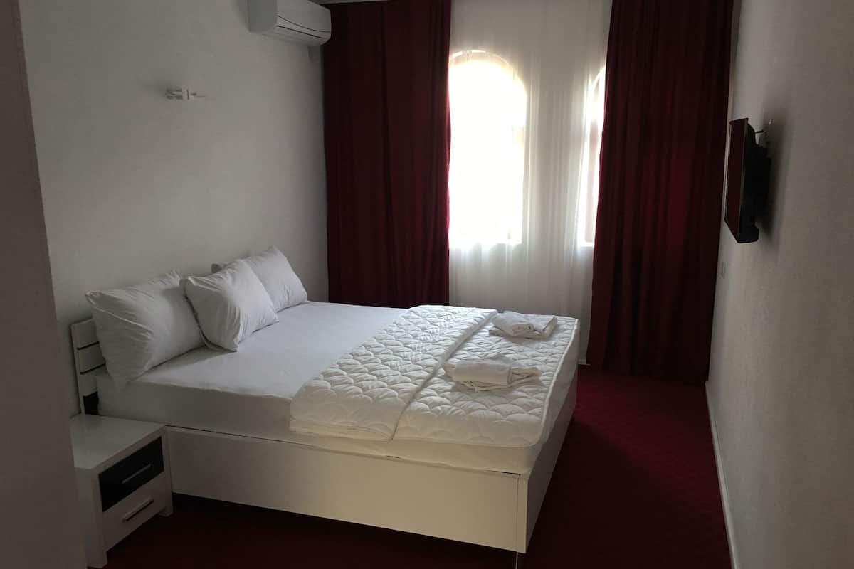 A neatly made bed with white linens and a quilt, a bedside table with drawers, and a wall-mounted air conditioning unit.