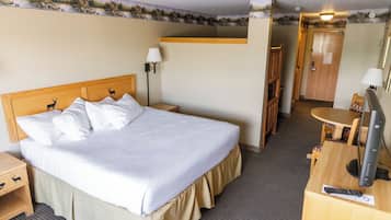 Quarto de hotel com uma cama, uma escrivaninha com computador e luminária, uma cadeira, uma mesa pequena e uma porta.