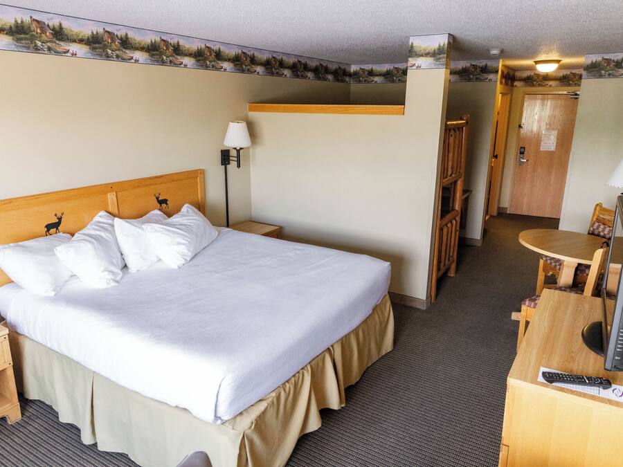 Quarto de hotel com uma cama, uma escrivaninha com computador e luminária, uma cadeira, uma mesa pequena e uma porta.