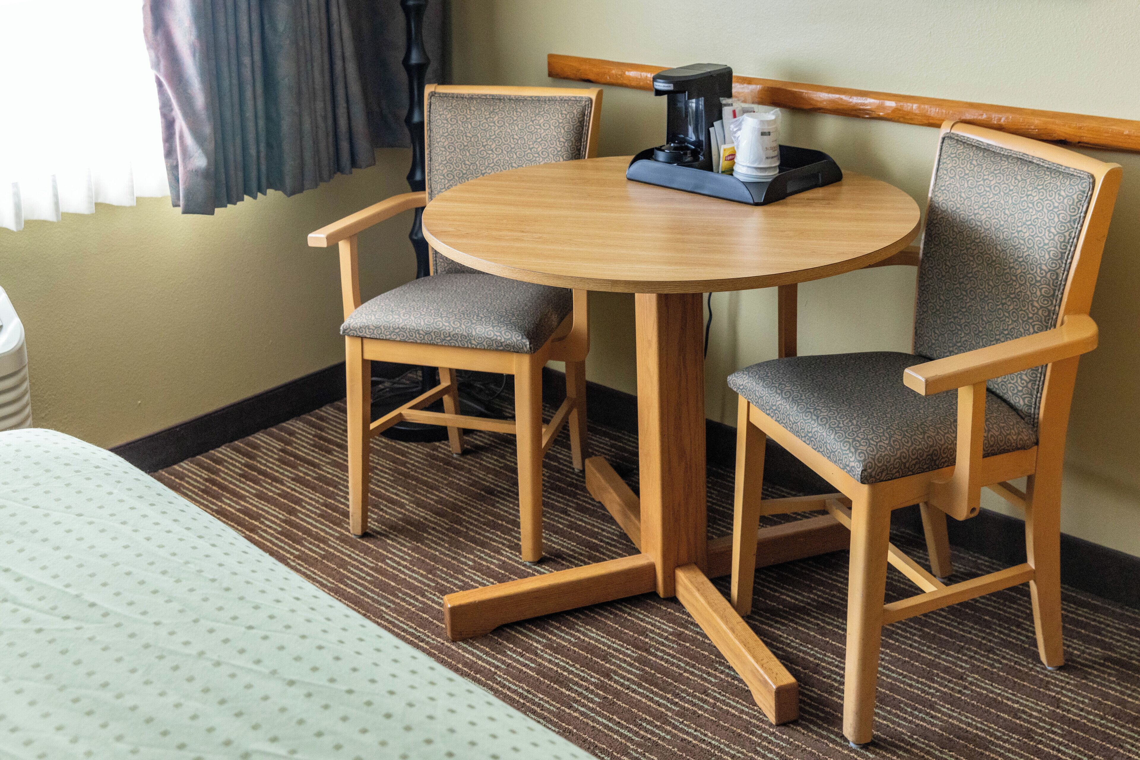 A hotel room with a round wooden table, two upholstered chairs, a coffee maker, and a bed with a patterned bedspread.