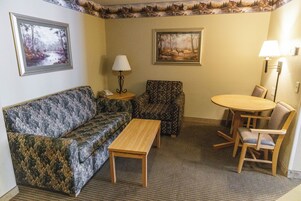 Un salón con un sofá, un sillón estampado, una mesa de centro de madera y una pequeña mesa redonda con dos sillas. Hay dos lámparas de pared y dos cuadros enmarcados sobre el sofá.