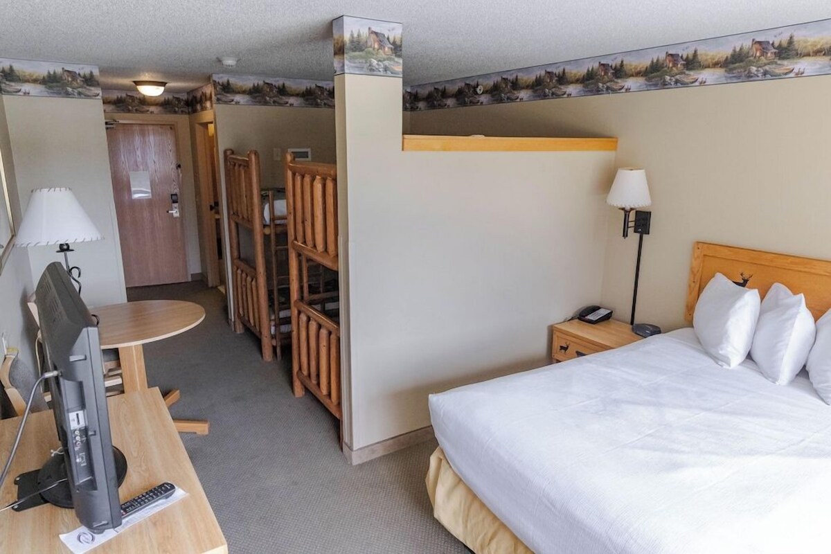 Habitación de hotel con cama, escritorio con computadora, silla, mesita pequeña y mesita de noche con lámpara.