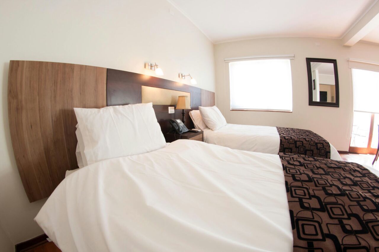 A hotel room with two beds, a wooden headboard, bedside lamps, a mirror, and a window with blinds.