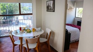 Une pièce avec une table à manger dressée pour deux personnes, un lit recouvert d’une couverture rouge et blanche, et une fenêtre donnant sur la rue.