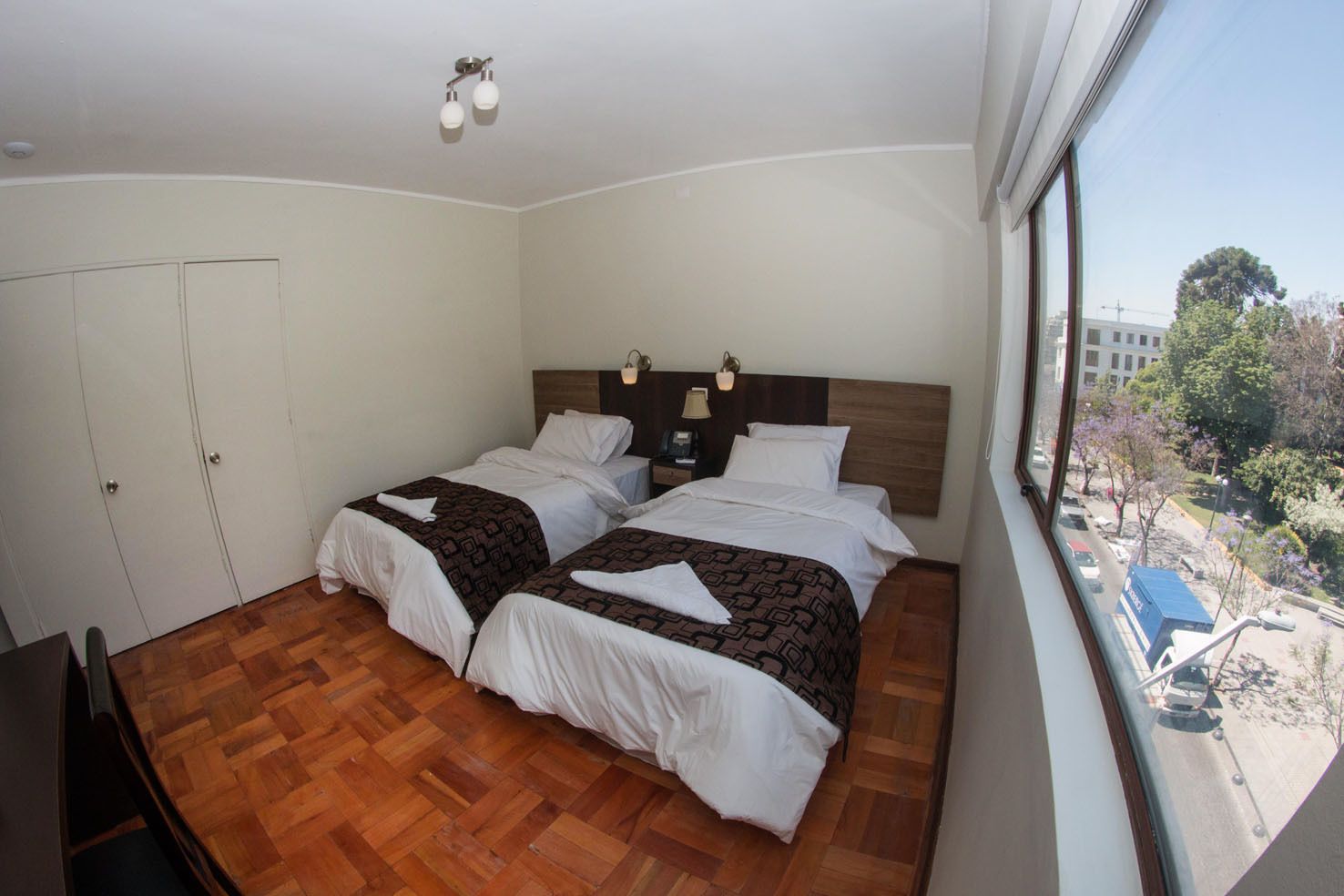 Habitación de hotel con dos camas, piso de madera, un ventanal con vistas a la ciudad y una lámpara en el techo.