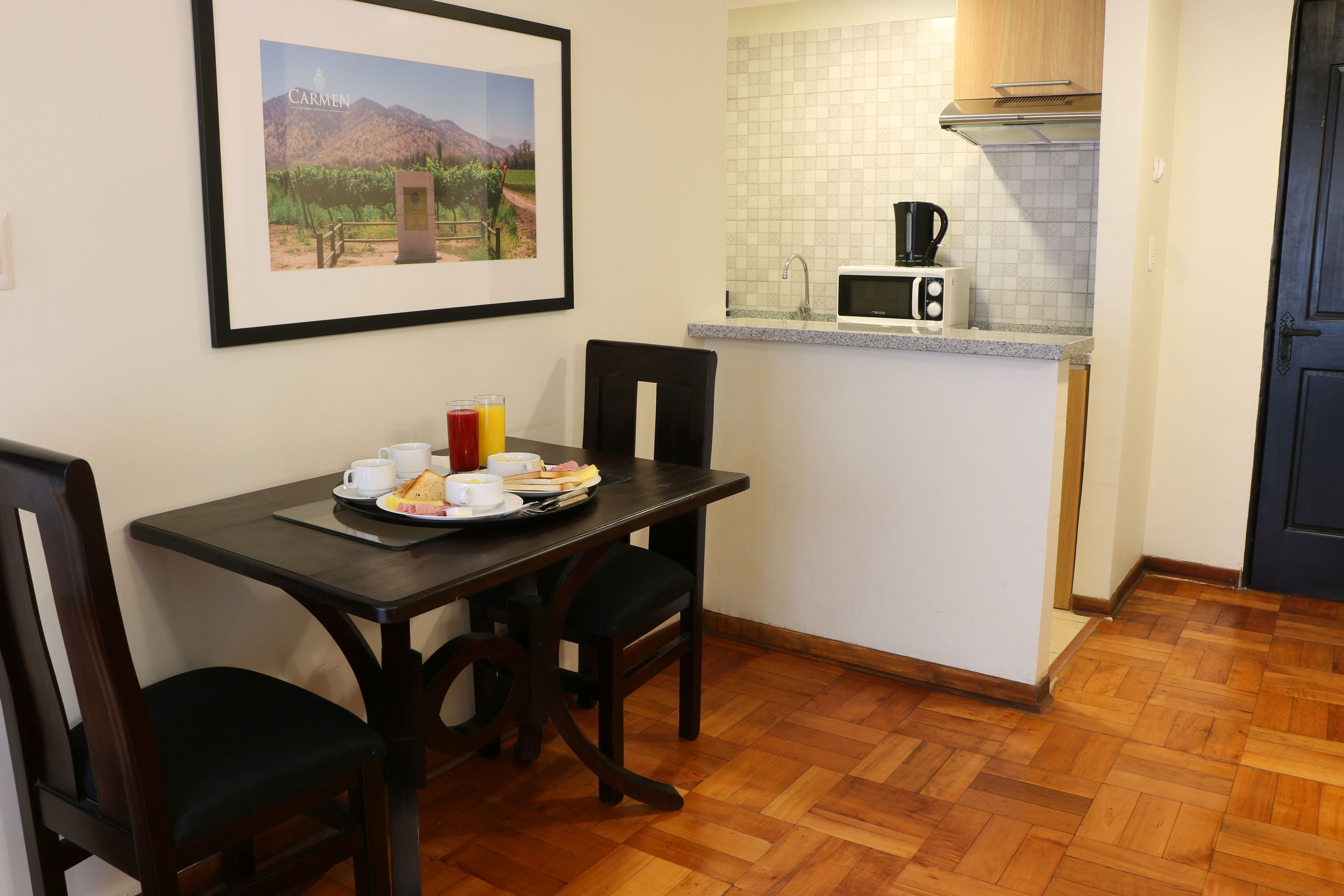 A dining area with a table set for two, a framed picture on the wall, and a kitchen area with a microwave and sink.