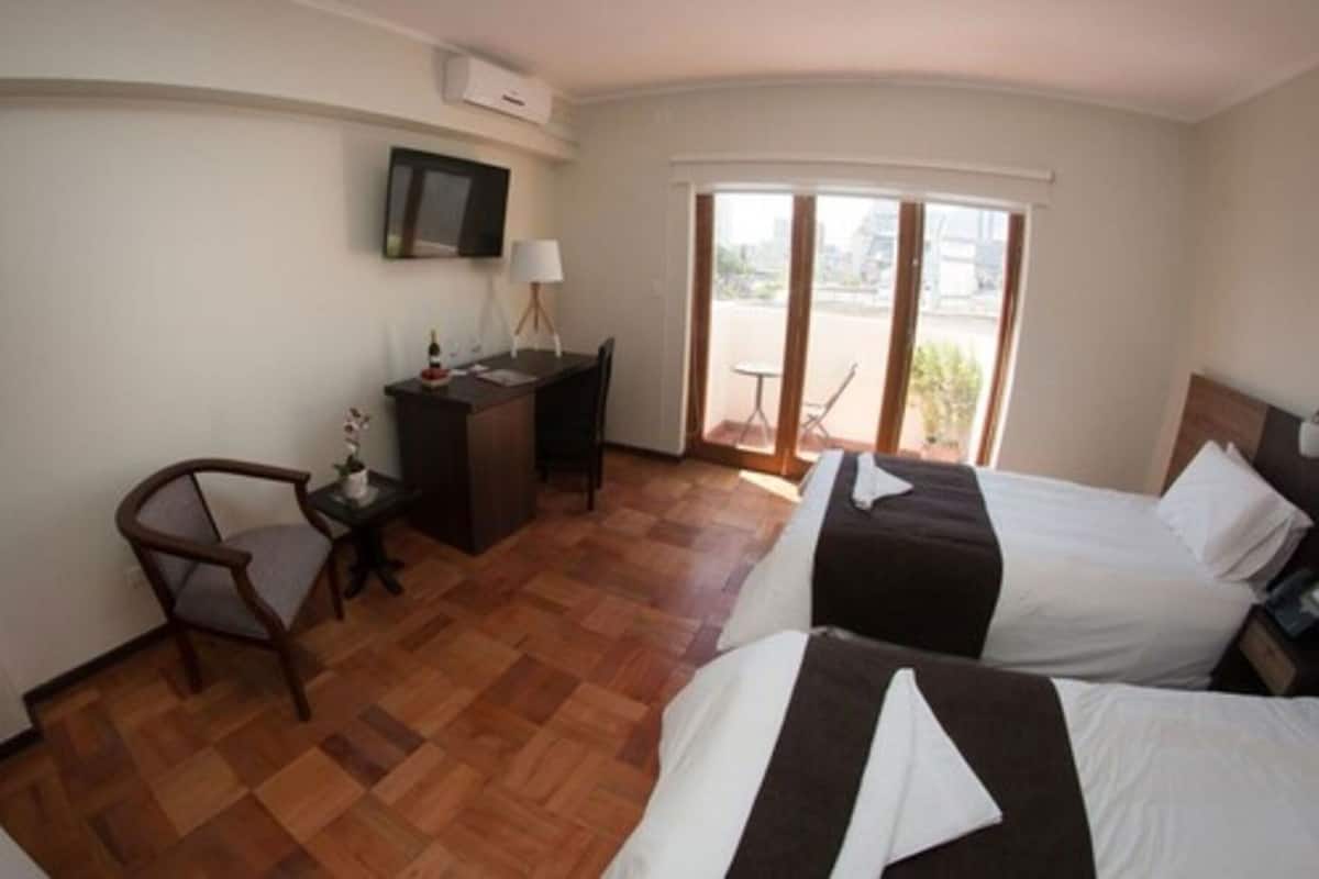 Une chambre d’hôtel avec un bureau, deux lits, une chaise et une grande fenêtre.