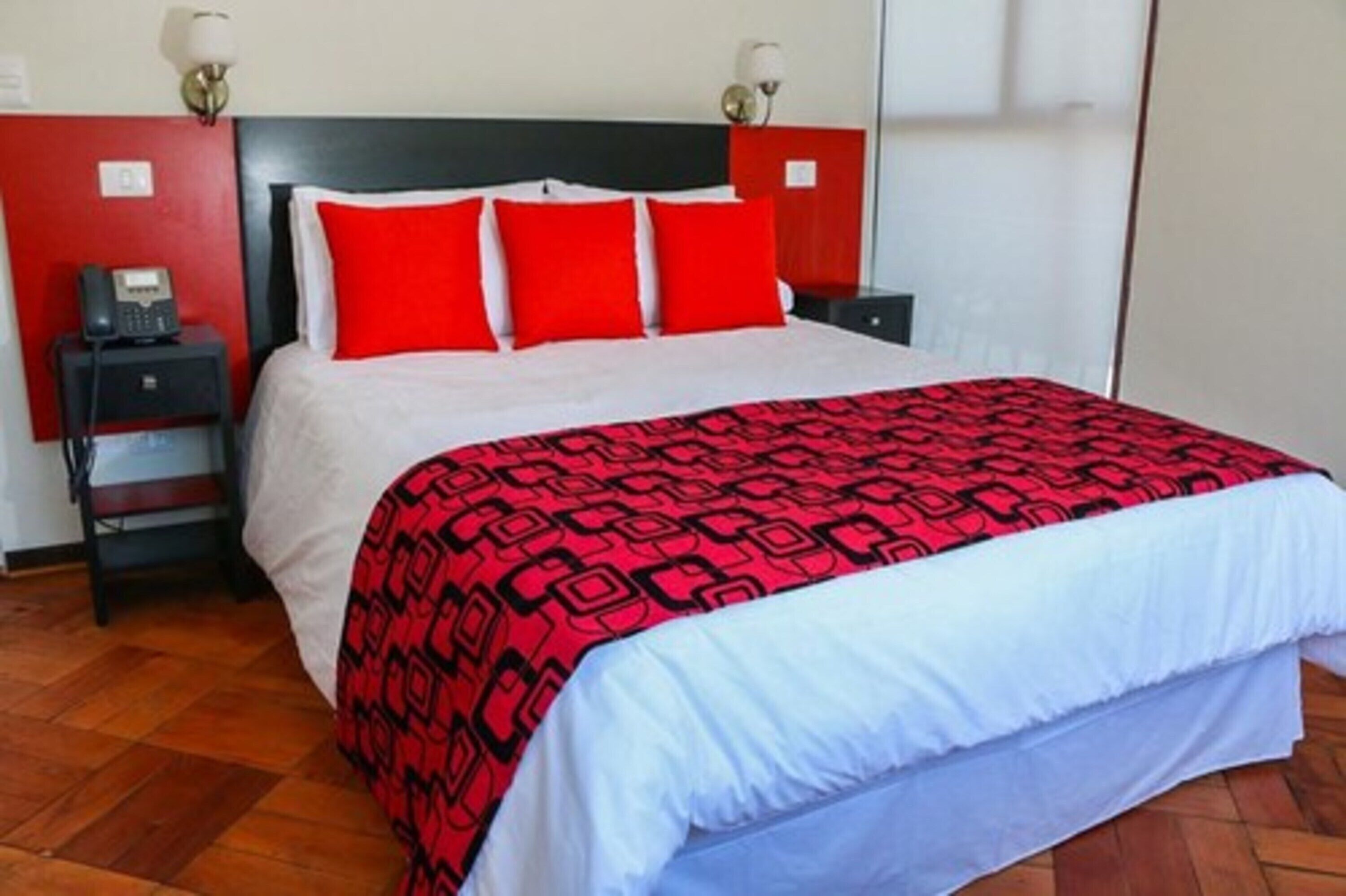 A hotel room with a bed, red and white bedding, a patterned throw, a bedside table with a lamp, and a telephone.