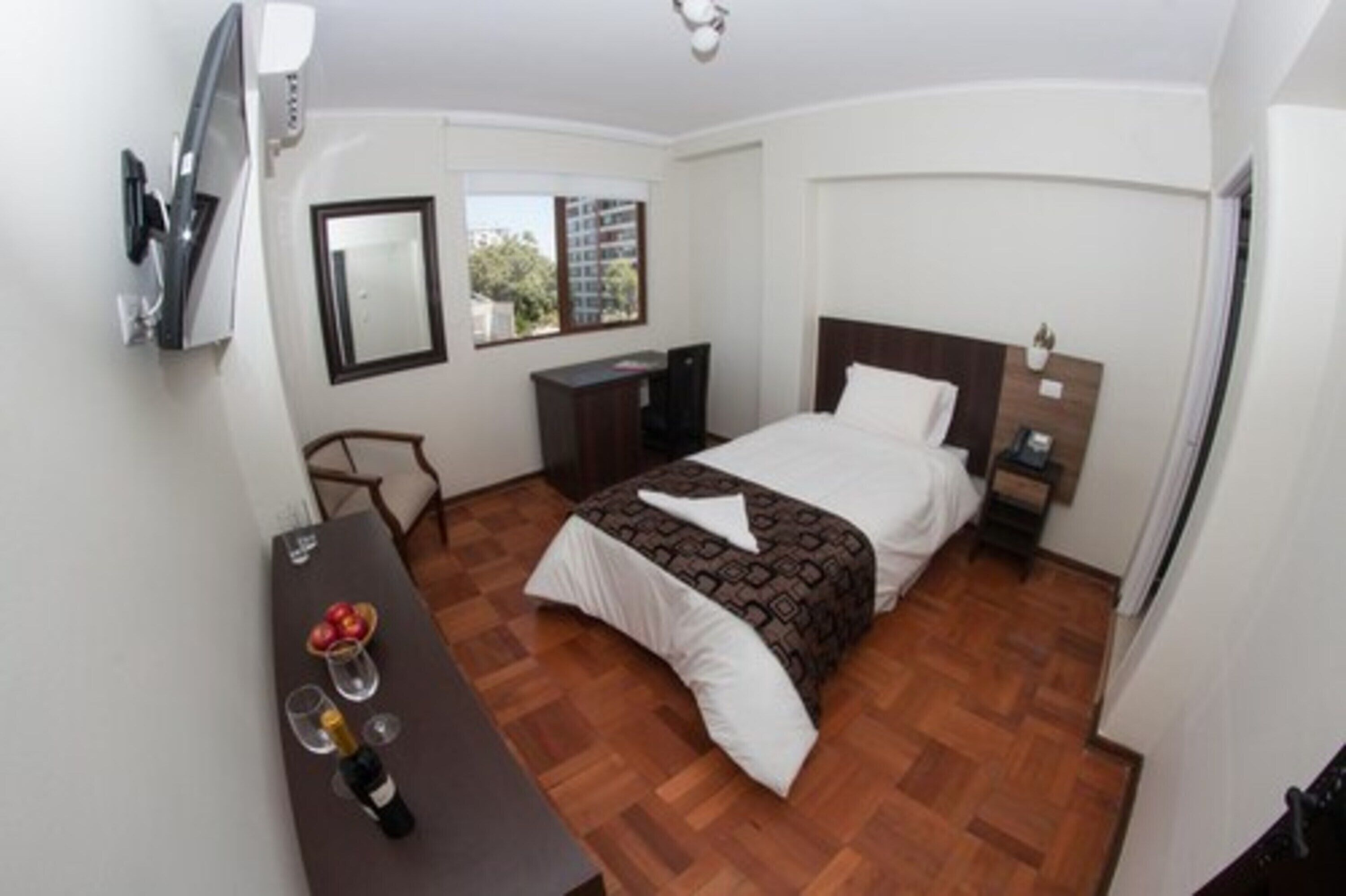 A bedroom with a bed, a desk with a bottle and glasses, a chair, a TV mounted on the wall, and a window with a view of buildings.